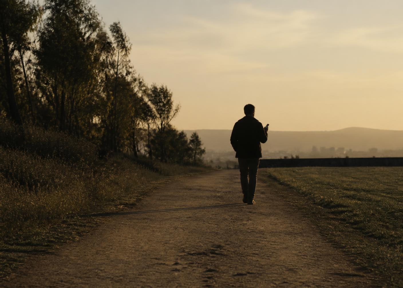 Persona caminando al atardecer por un sendero, con el celular en la mano.